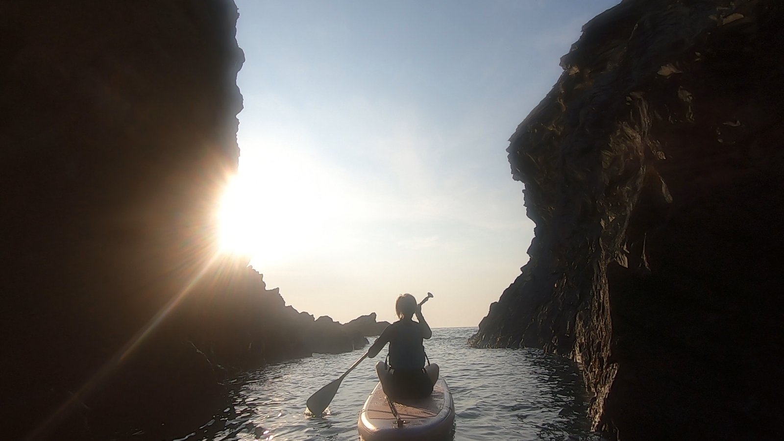 A paddleboarder gliding between towering cliffs toward the low evening sun at the mouth of the Witches Cauldron, Pembrokeshire.